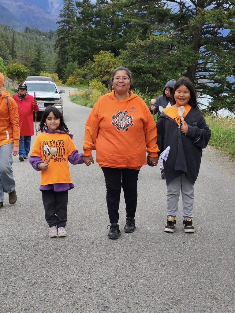 Three women walk hand-in-hand along a road, wearing orange clothing with "Every Child Matters" slogans